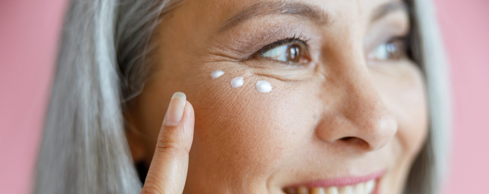 Beautiful mature Asian lady applies antiaging cream under eye on pink background